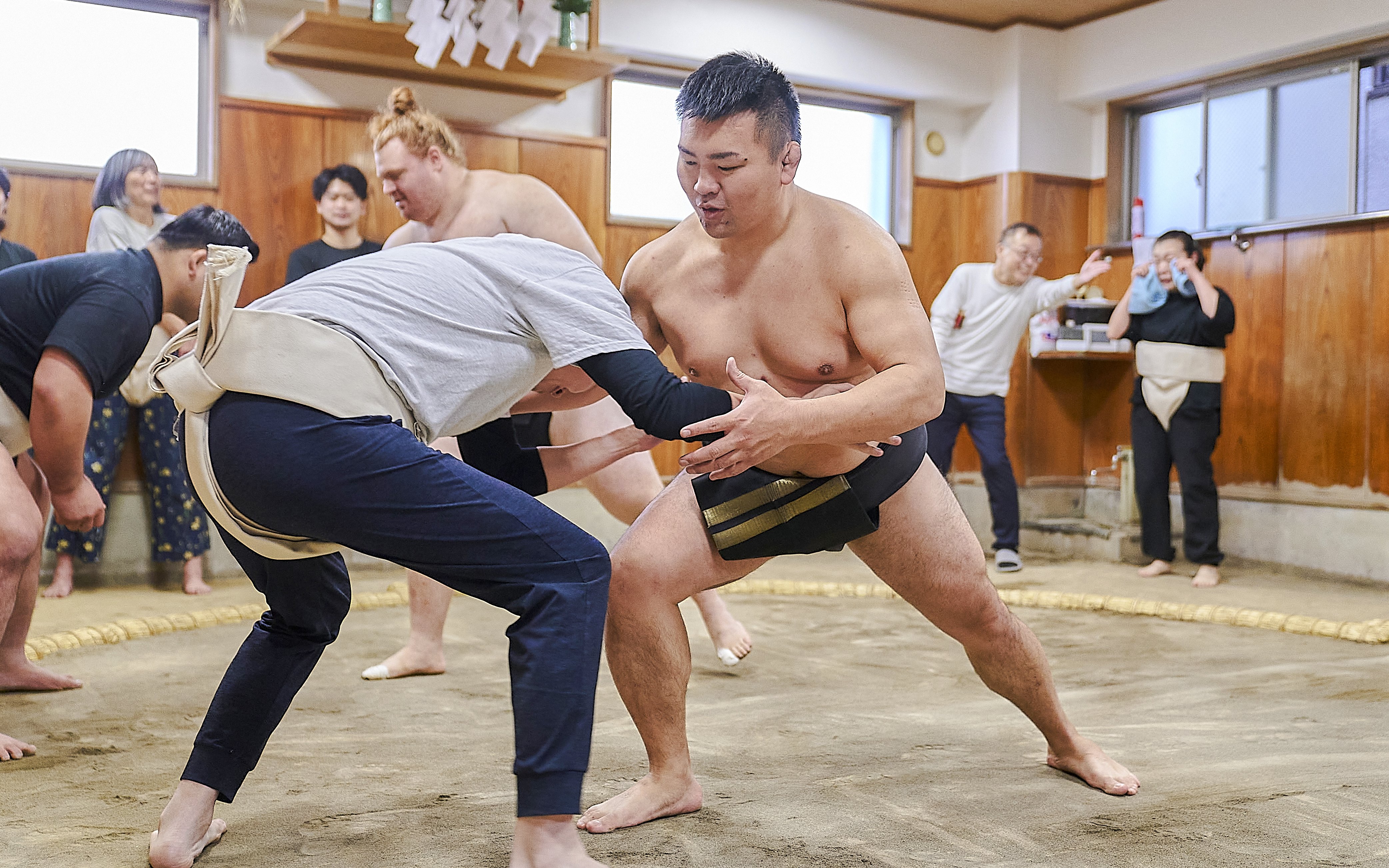 Sumo wrestlers practicing in a Koto City dojo with spectators watching.