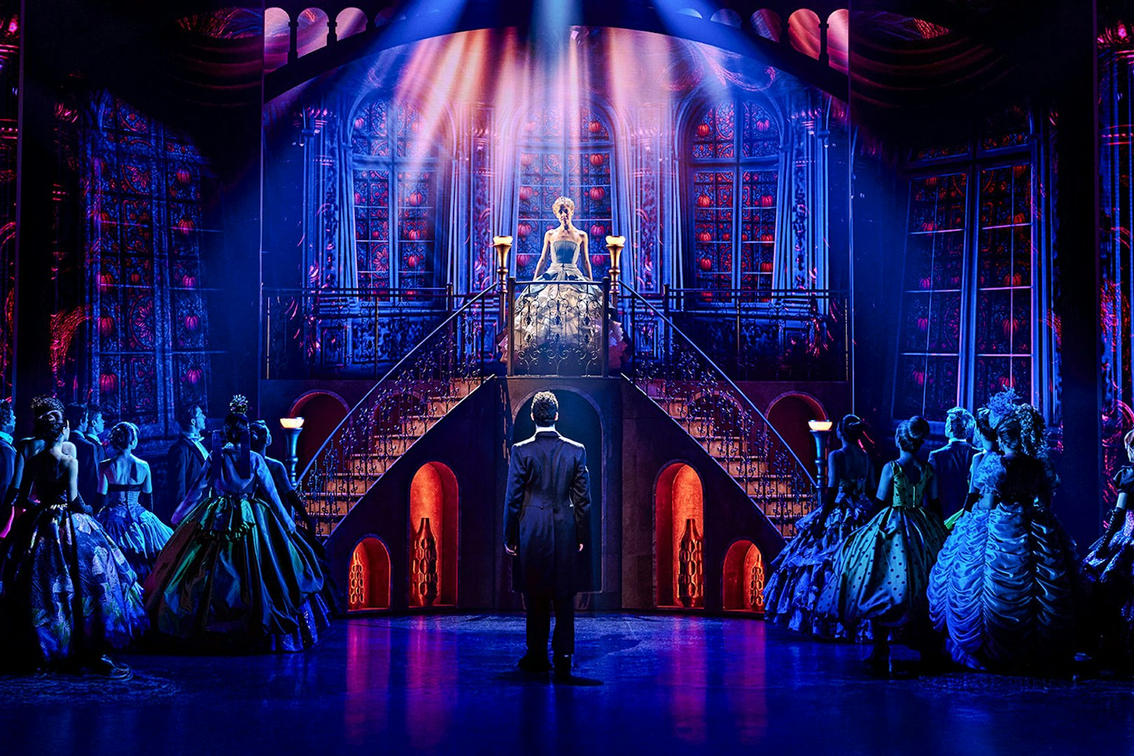 Cinderella on stage at Madrid theater, standing on staircase, surrounded by cast in elegant costumes.