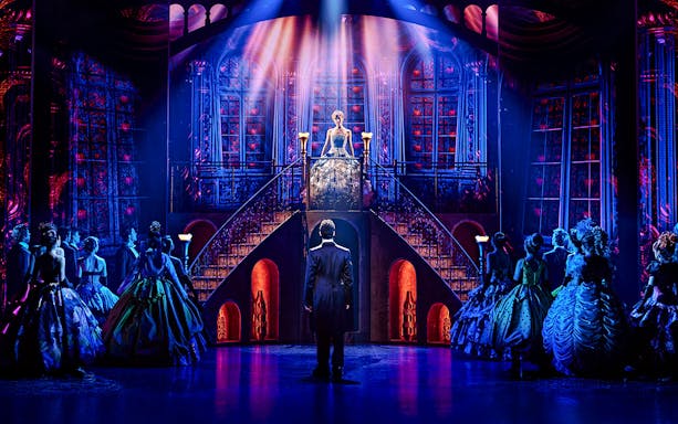Cinderella on stage at Madrid theater, standing on staircase, surrounded by cast in elegant costumes.