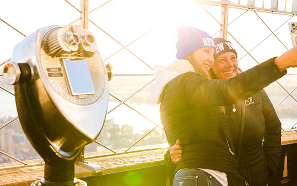 Guests taking a selfie on the Empire State Building's observation deck with a viewfinder nearby.