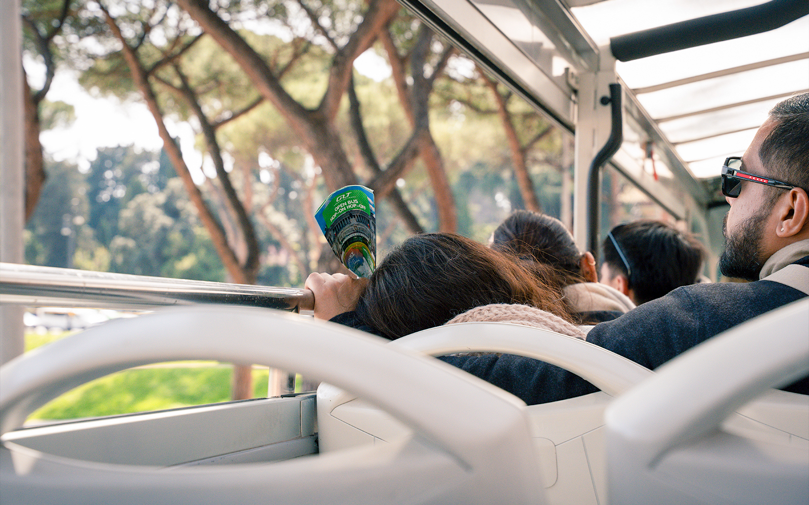 Passengers on Rome hop-on hop-off bus tour holding a brochure, surrounded by trees.