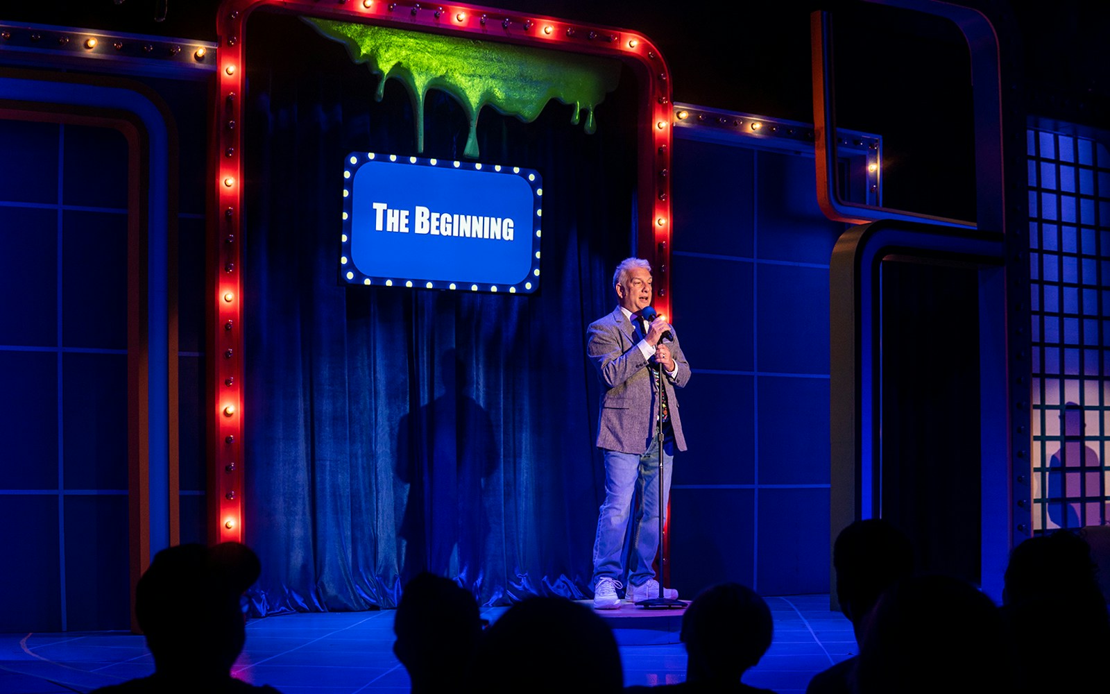 Man on stage with microphone, blue curtain backdrop, sign reads "The Beginning," related to "The Life and Slimes of Marc Summers.