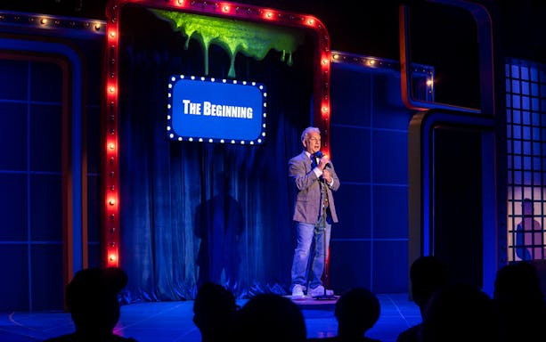 Man on stage with microphone, blue curtain backdrop, sign reads "The Beginning," related to "The Life and Slimes of Marc Summers.