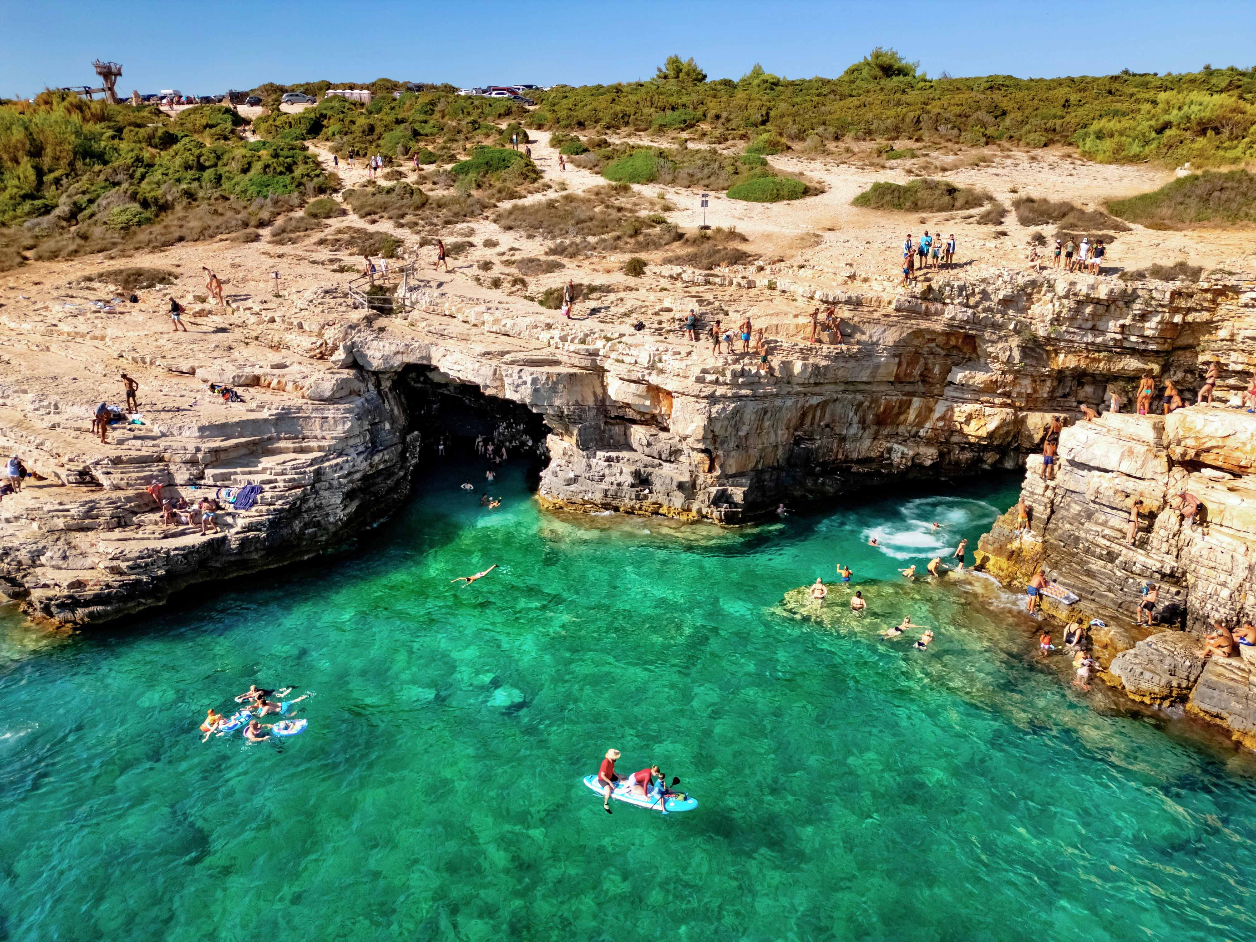 Visitors swimming and paddleboarding near the rocky entrance of Green Cave, Croatia.