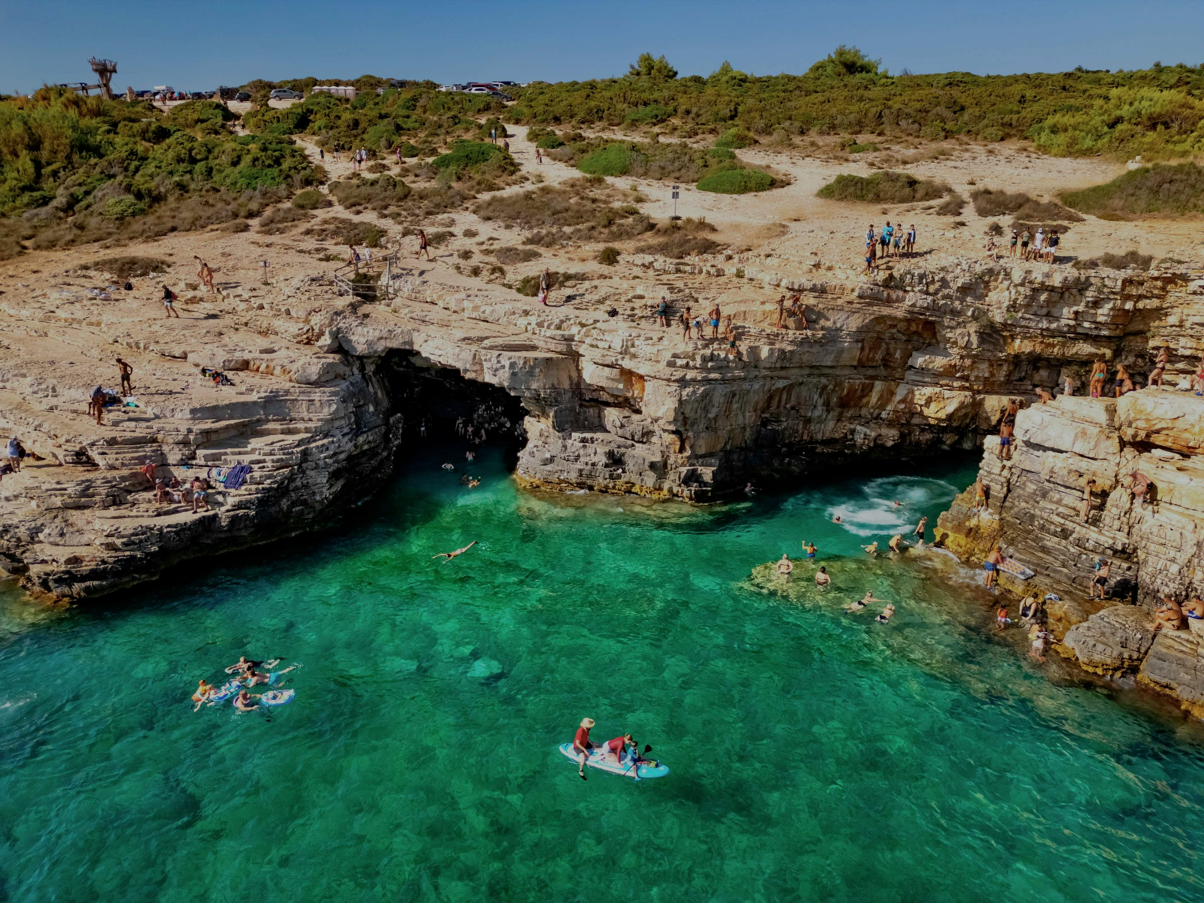 Visitors swimming and paddleboarding near the rocky entrance of Green Cave, Croatia.