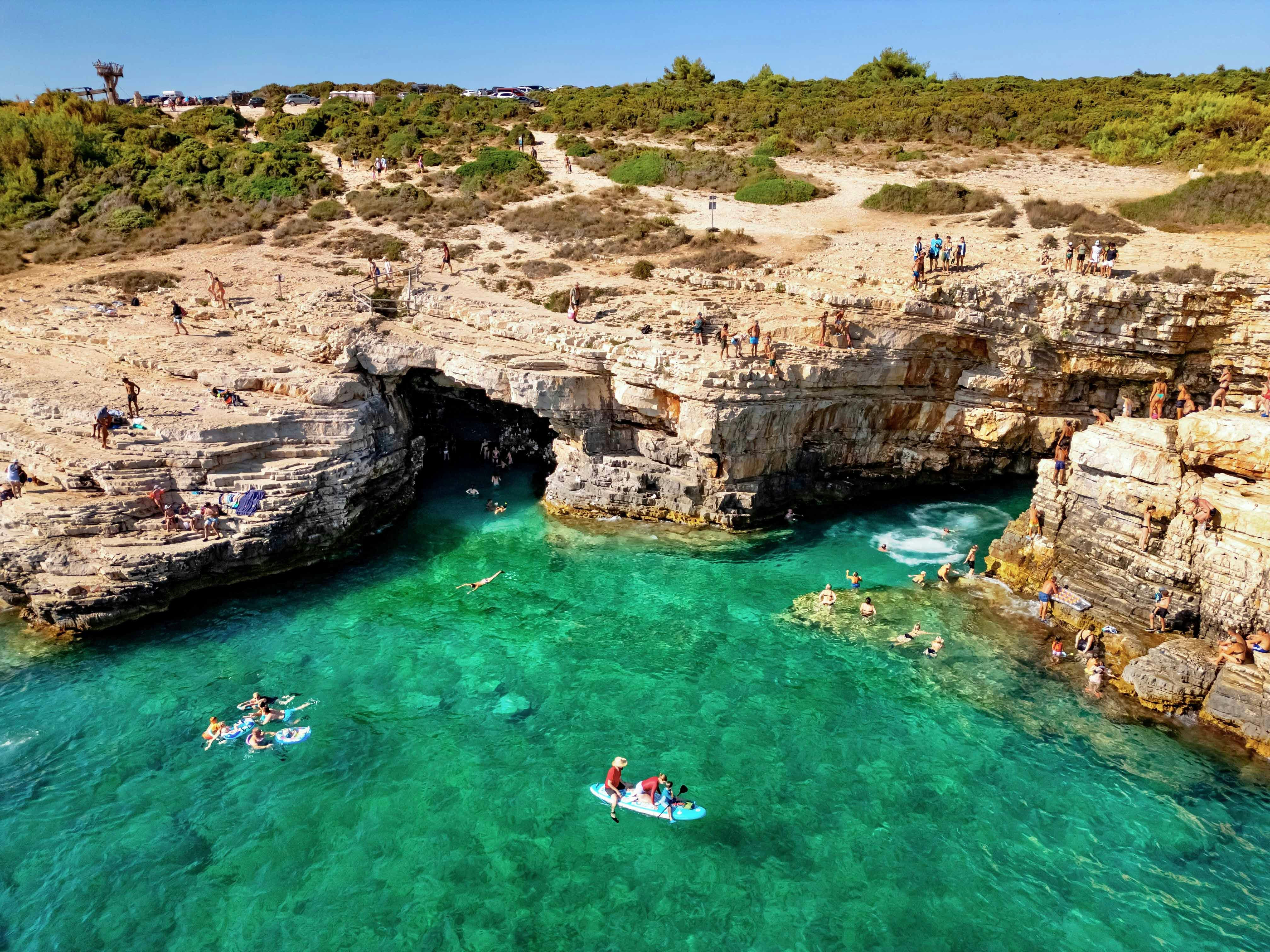 Visitors swimming and paddleboarding near the rocky entrance of Green Cave, Croatia.