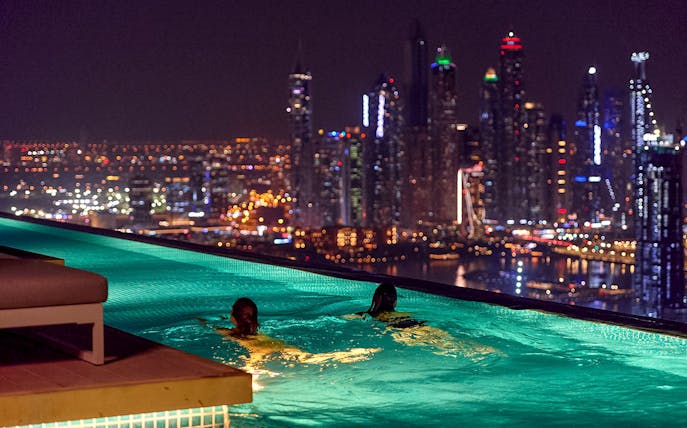 Tourists swimming at Aura Sky Pool with Dubai skyline at night.