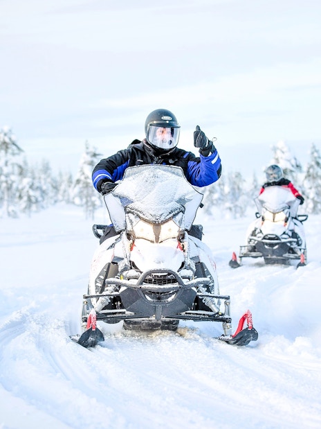 Snowmobile riders on a snowy trail in Rovaniemi, surrounded by snow-covered trees.