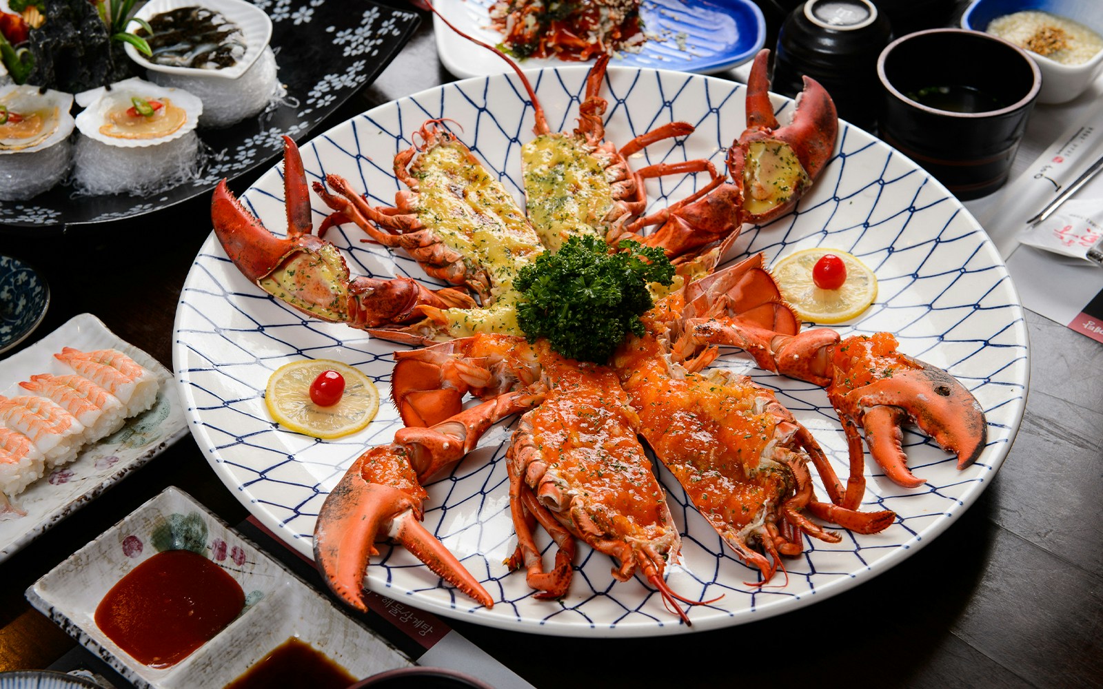 Lobster platter with lemon slices and garnish, surrounded by sushi and dipping sauces.