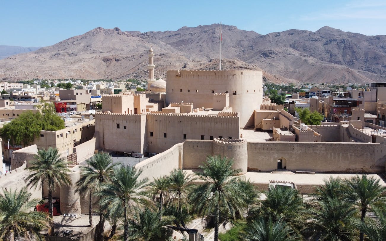 Nizwa Fort in Oman with surrounding palm trees and mountains in the background.