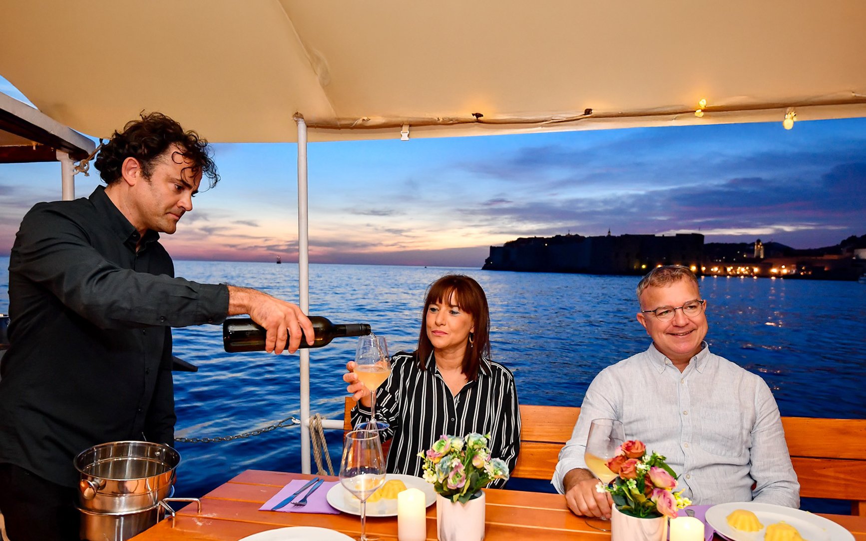 Couple enjoying drinks on Dubrovnik sunset cruise with waiter pouring wine.