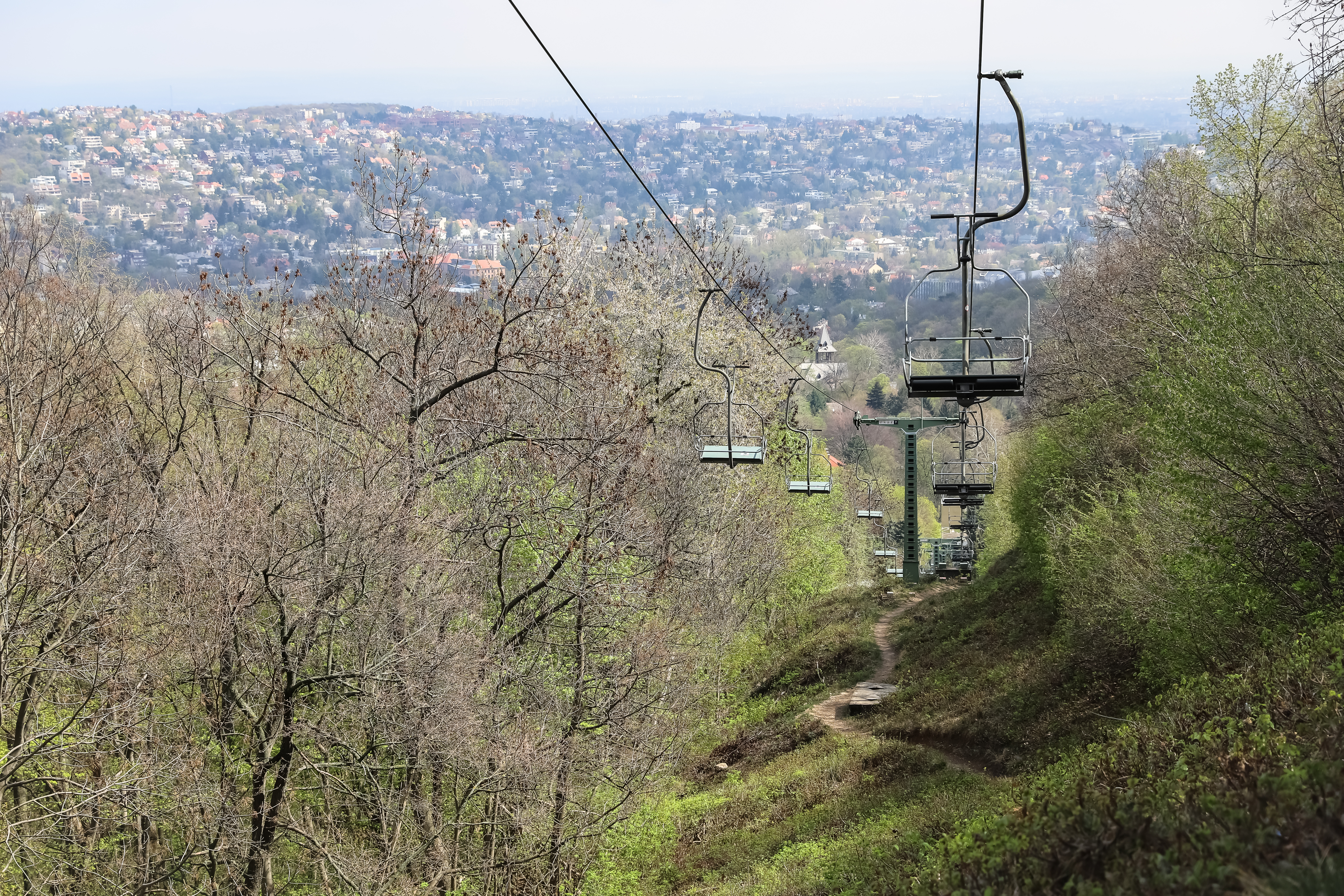 Chairlift ascending over a forested hillside with a cityscape in the background.