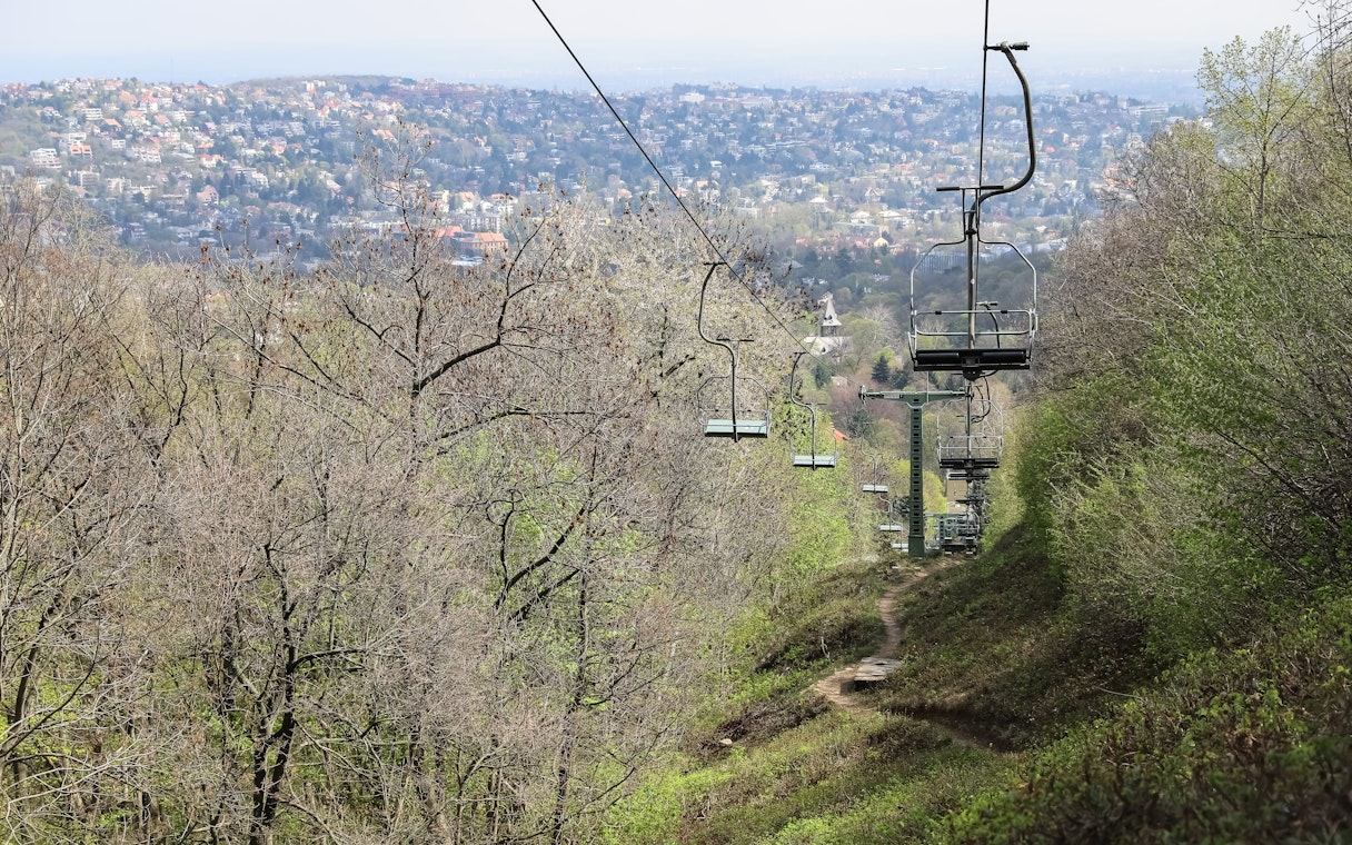 Chairlift ascending over a forested hillside with a cityscape in the background.