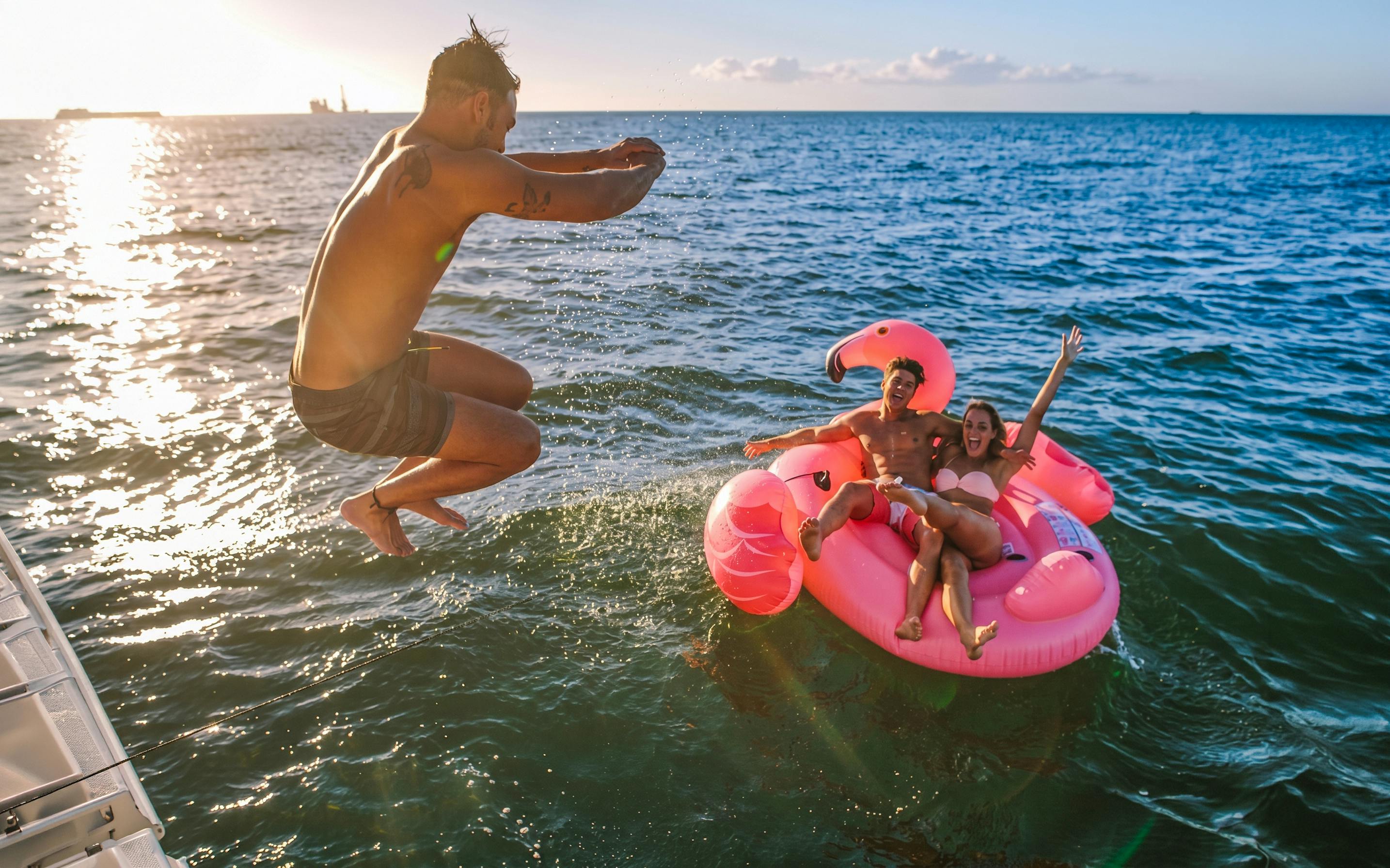 Man jumping into the sea near a pink flamingo float with two people on a Heraklion catamaran cruise.