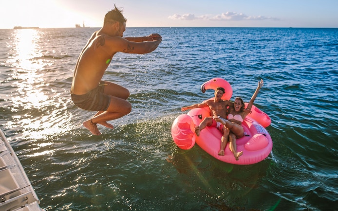 Man jumping into the sea near a pink flamingo float with two people on a Heraklion catamaran cruise.
