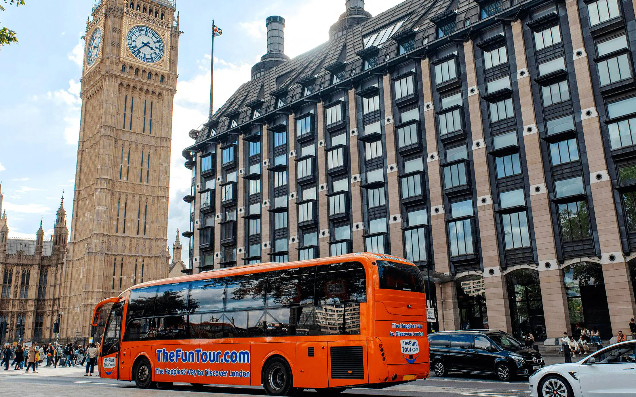 Orange tour bus near Big Ben in London, promoting The Fun Tour with live theatre, comedy, and music.
