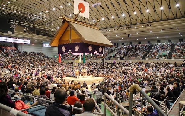 Sumo wrestlers competing in a packed Tokyo arena under a traditional Japanese roof.