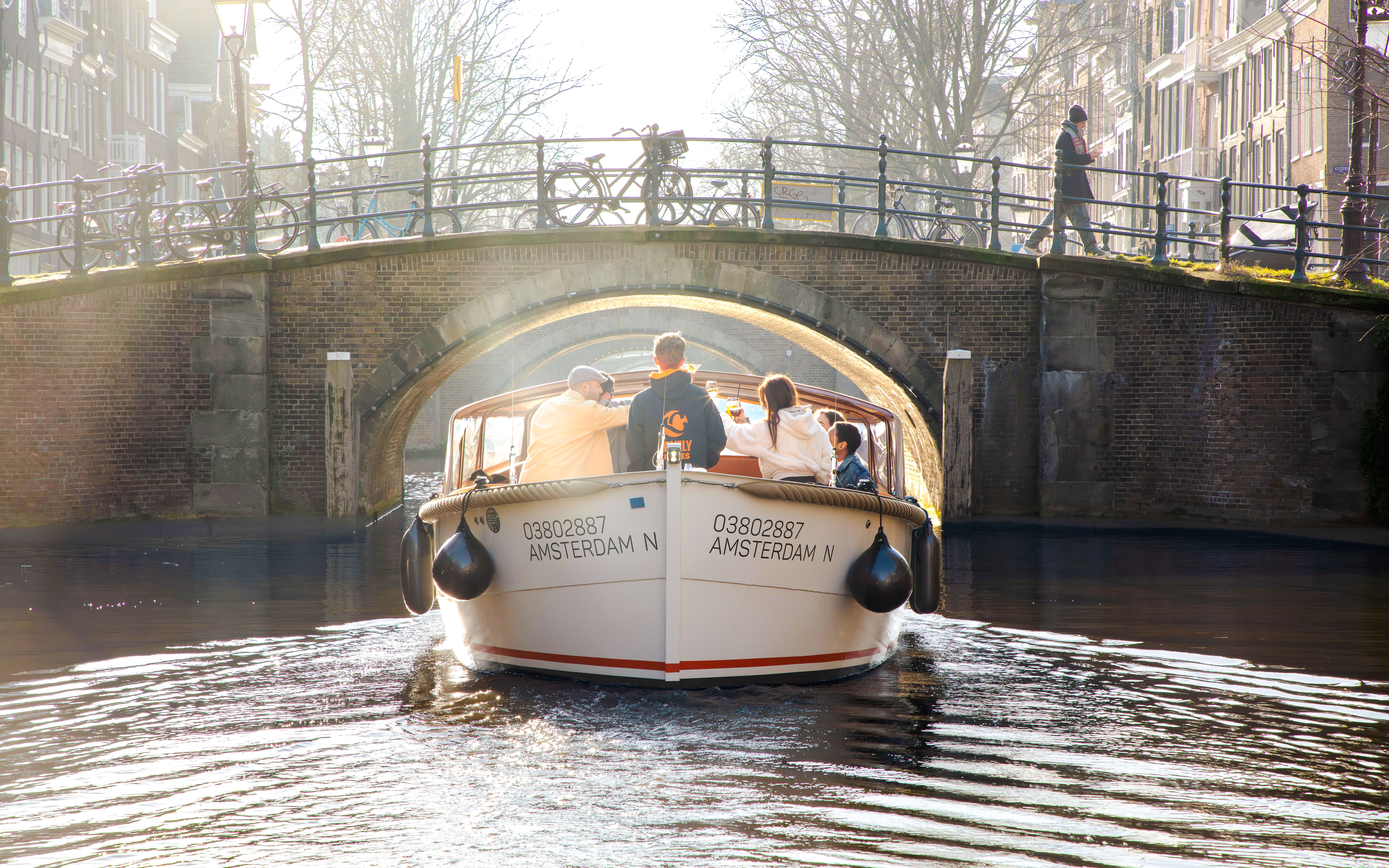 Open boat cruise under a canal bridge in Amsterdam with passengers enjoying the view.