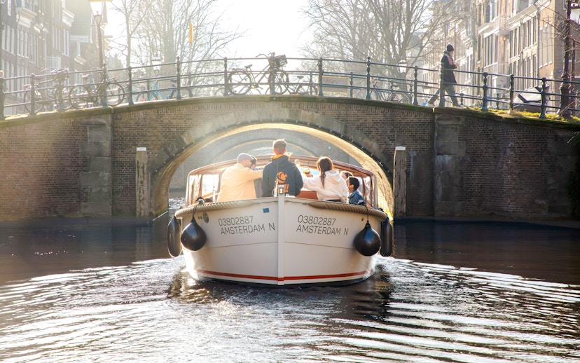 Open boat cruise under a canal bridge in Amsterdam with passengers enjoying the view.