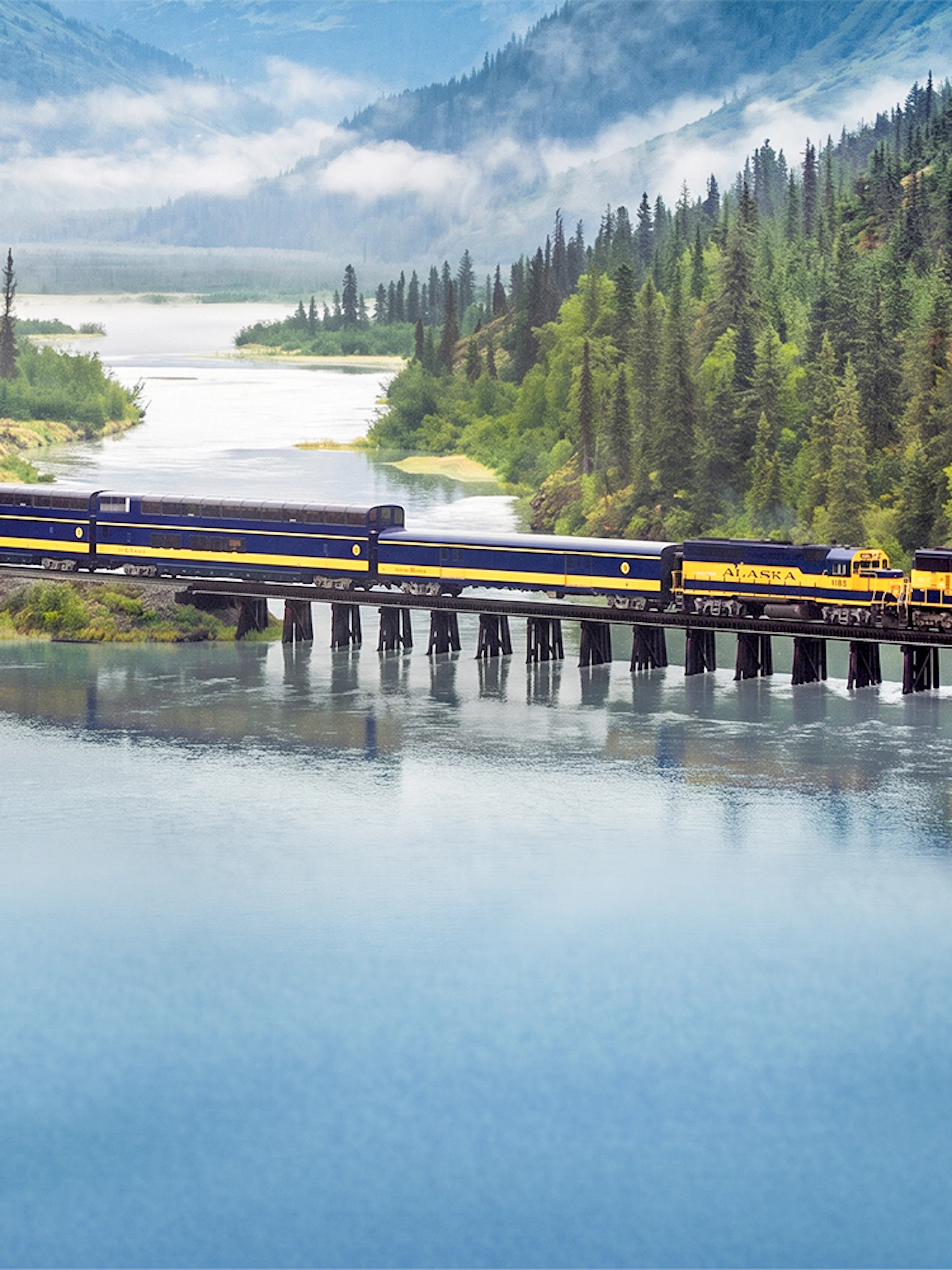Alaska Railroad train crossing a bridge over a scenic river with forested mountains.