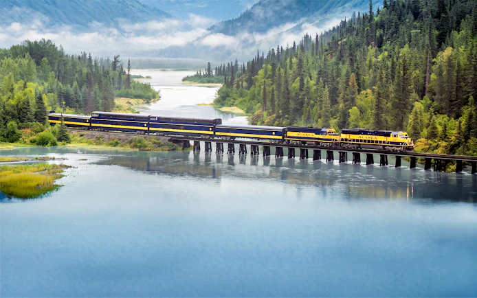 Alaska Railroad train crossing a bridge over a scenic river with forested mountains.