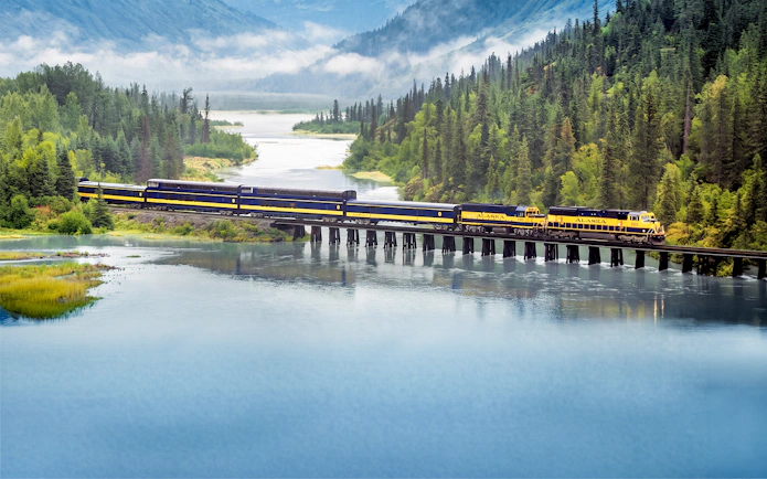 Alaska Railroad train crossing a bridge over a scenic river with forested mountains.
