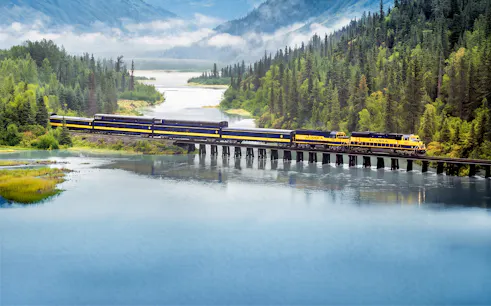 Alaska Railroad train crossing a bridge over a scenic river with forested mountains.