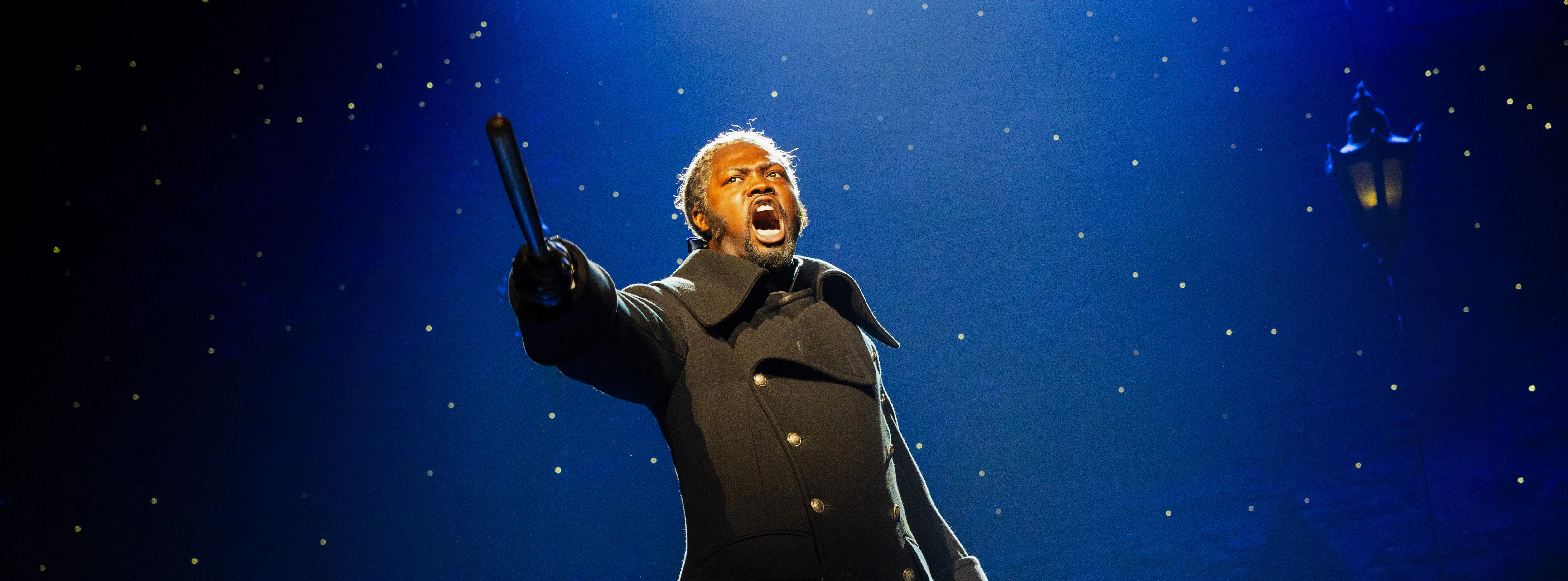 Performer on stage during Les Misérables musical, holding a hat and pointing dramatically.