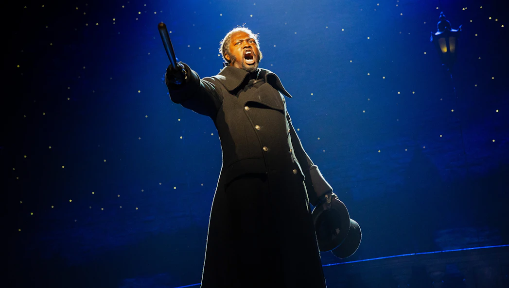 Performer on stage during Les Misérables musical, holding a hat and pointing dramatically.