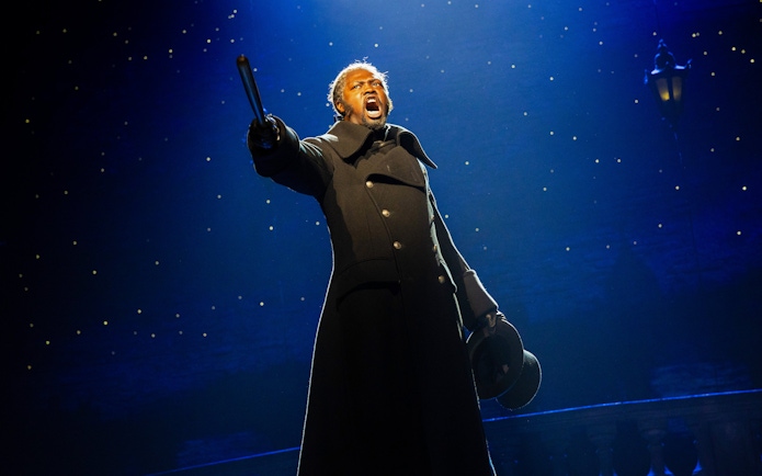 Performer on stage during Les Misérables musical, holding a hat and pointing dramatically.