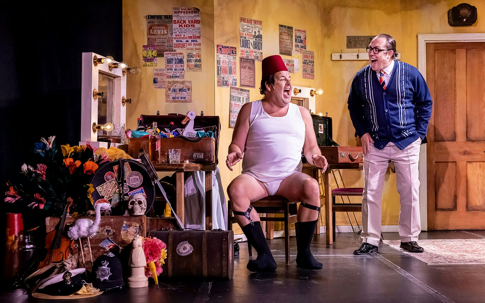 Two actors in a comedic scene on stage, surrounded by props and vintage posters, performing "The Last Laugh.