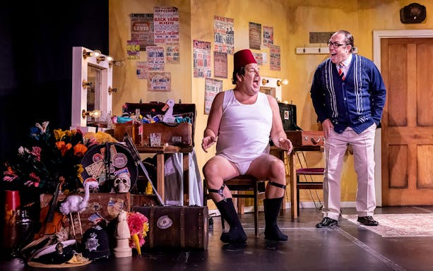 Two actors in a comedic scene on stage, surrounded by props and vintage posters, performing "The Last Laugh.