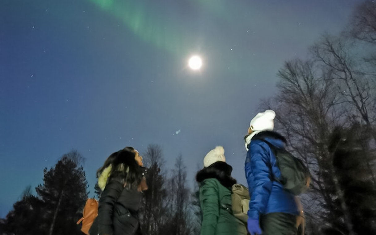 Friends watching northern lights in Lapland under a moonlit sky.