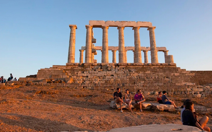 Temple of Poseidon at sunset with visitors seated on ancient stones.