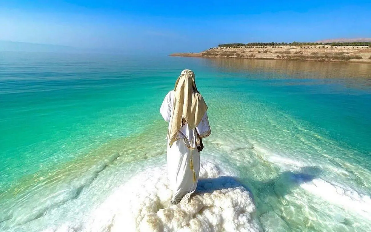 Person standing on salt formations at the Dead Sea, Jordan, with clear blue water.