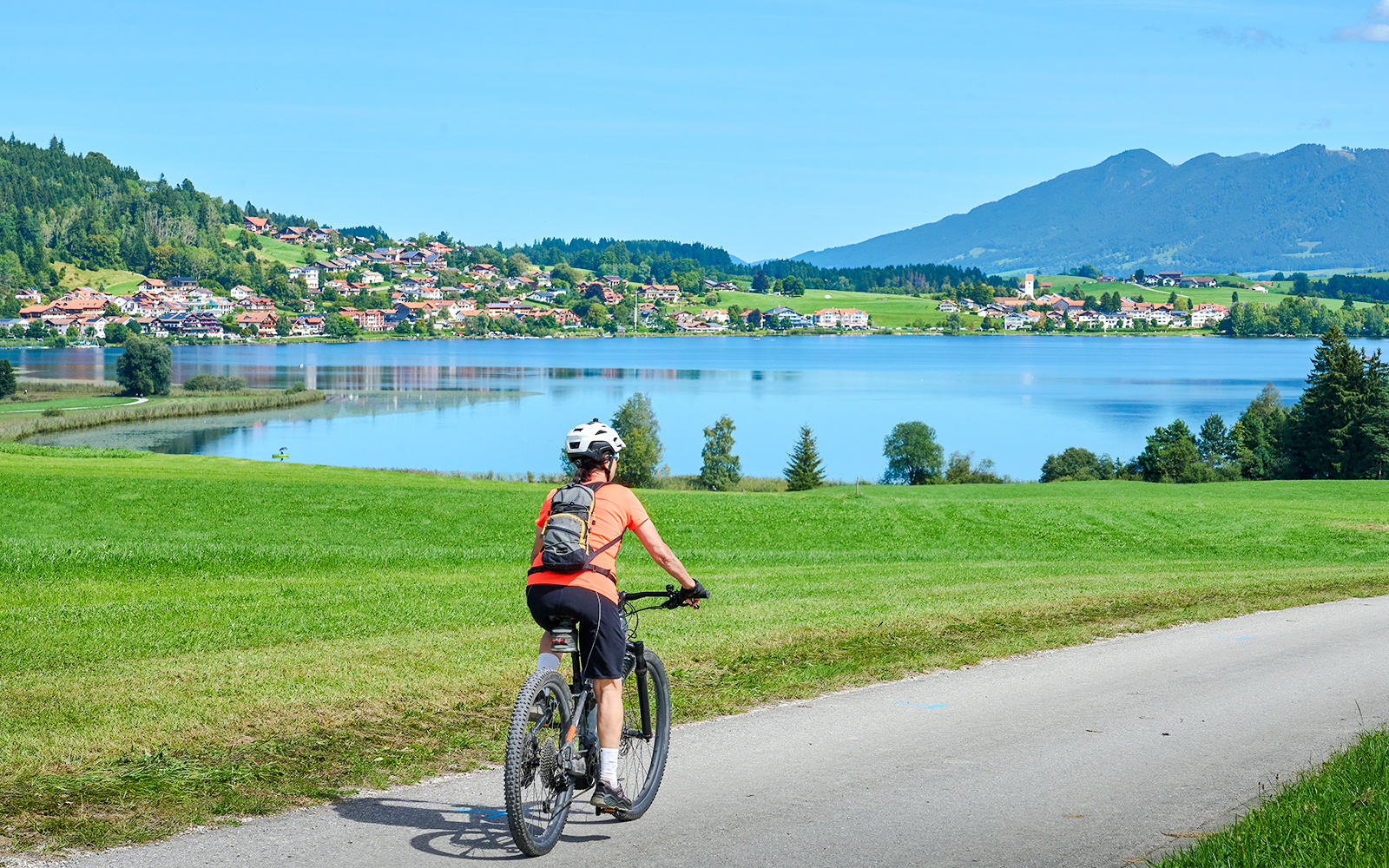 Cyclist riding along Alpsee with scenic view of lake and mountains.