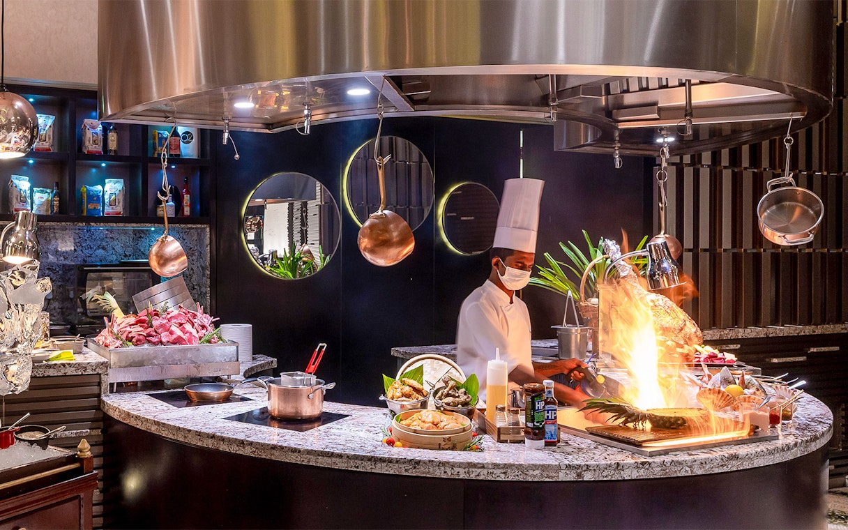 Chef preparing buffet dishes at Mundo Jumeirah Emirates Towers with open flame cooking.
