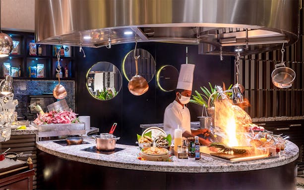 Chef preparing buffet dishes at Mundo Jumeirah Emirates Towers with open flame cooking.