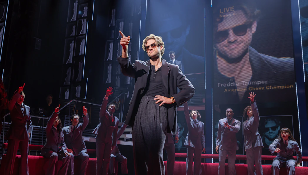 Stage scene from Chess Broadway show with performers in suits and large screen backdrop.