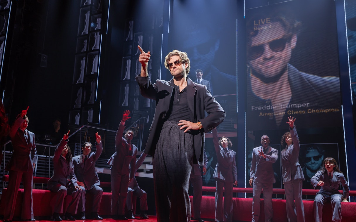 Stage scene from Chess Broadway show with performers in suits and large screen backdrop.