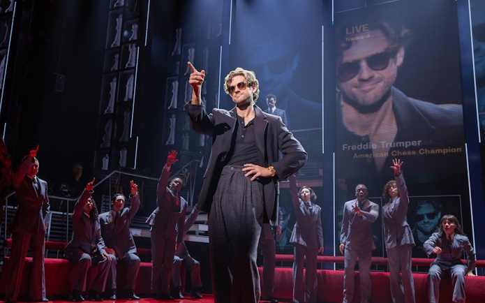 Stage scene from Chess Broadway show with performers in suits and large screen backdrop.