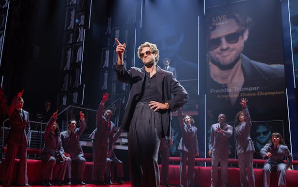 Stage scene from Chess Broadway show with performers in suits and large screen backdrop.