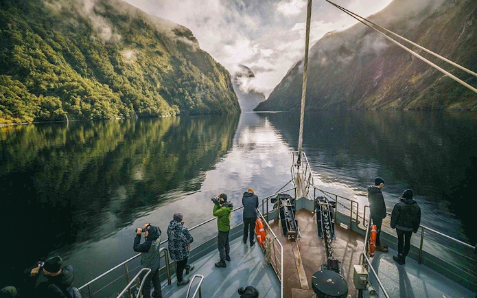 Tourists on a boat cruise through Doubtful Sound, surrounded by lush green cliffs and calm waters.