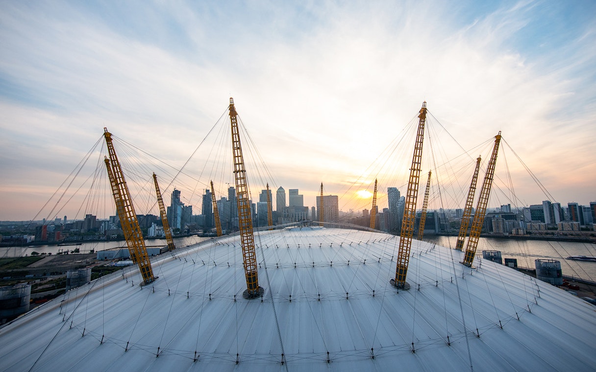 Climbers on the O2 Arena roof in London with city skyline at sunset.