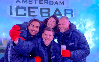 Group enjoying drinks at Icebar Amsterdam, wearing jackets and gloves, with ice wall backdrop.