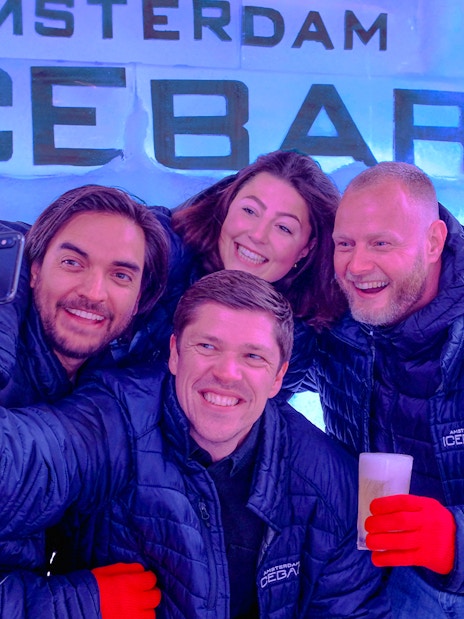 Group enjoying drinks at Icebar Amsterdam, wearing jackets and gloves, with ice wall backdrop.