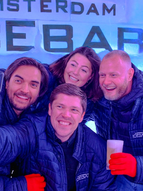 Group enjoying drinks at Icebar Amsterdam, wearing jackets and gloves, with ice wall backdrop.