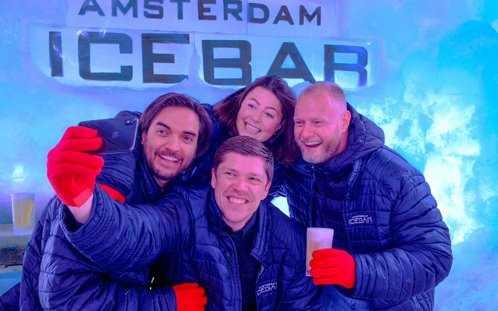 Group enjoying drinks at Icebar Amsterdam, wearing jackets and gloves, with ice wall backdrop.