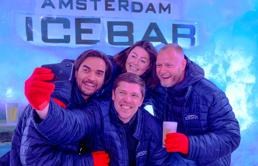 Group enjoying drinks at Icebar Amsterdam, wearing jackets and gloves, with ice wall backdrop.