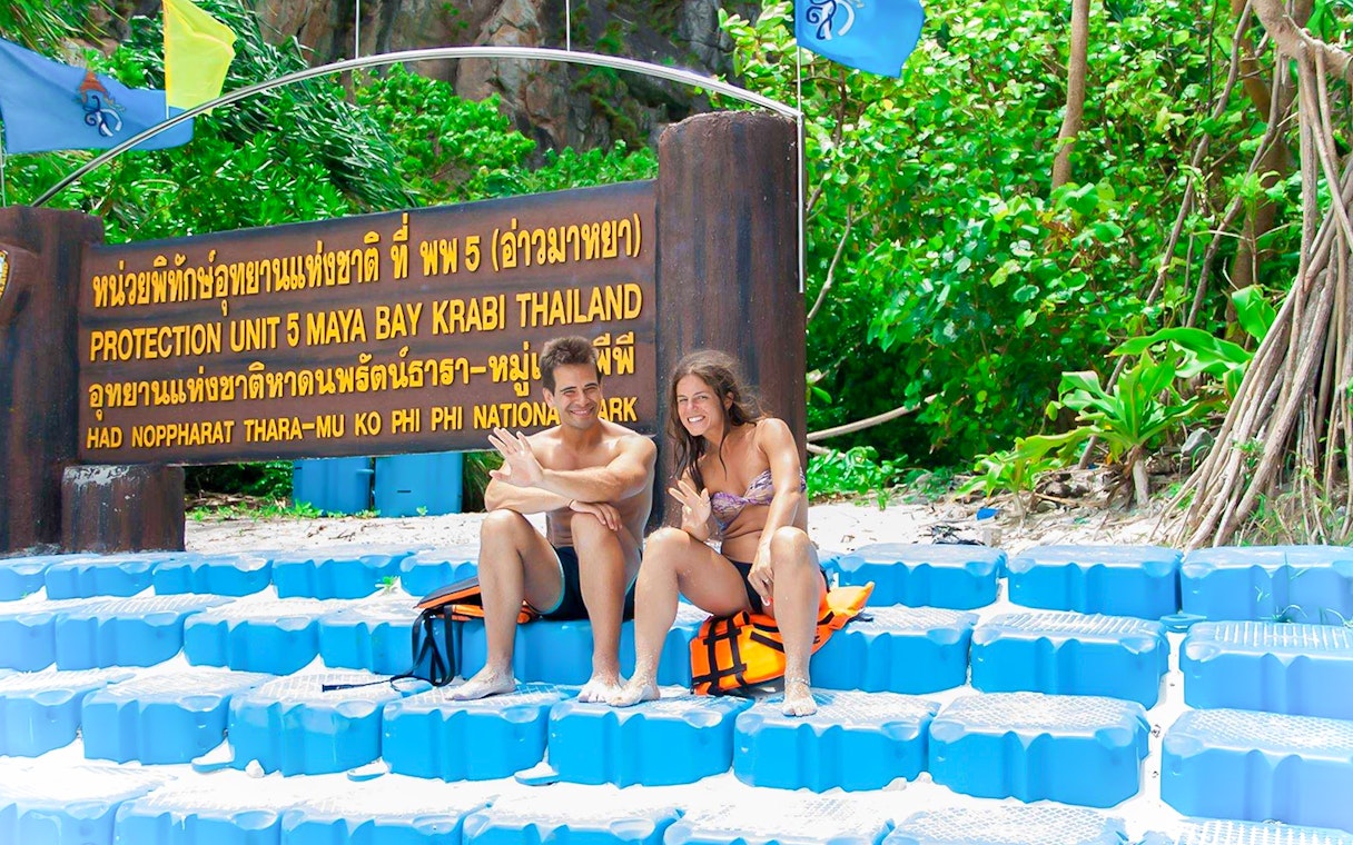 Couple sitting by Maya Bay sign, Phi Phi National Park, Thailand.